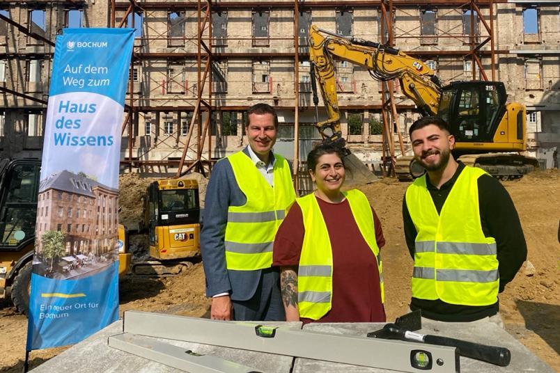 Drei Personen in Warnwesten auf einer Baustelle mit Hammer und Wasserwaage