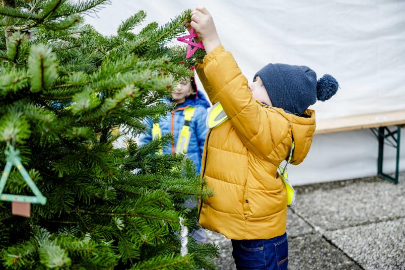 Kind in gelber jacke und mit blauer Mütze schmückt Tannenbaum.