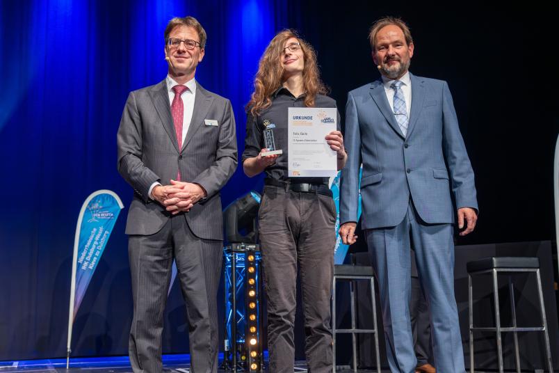 Drei Personen, einer davon mit Urkunde in der Hand