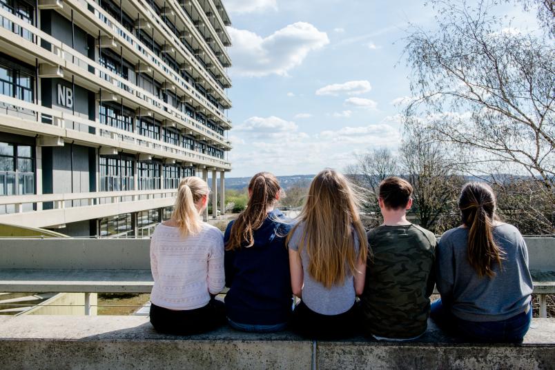 Fünf Personen sitzen mit dem Rücken zur Camera an einem Gebäude der RUB und Blicken in das Tal dahinter.