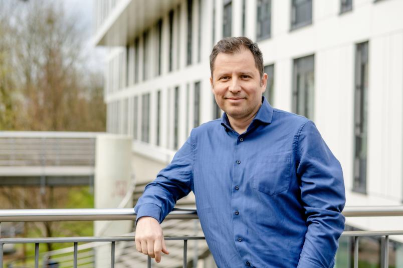 Porträtbild eines männlichen Forschers, der kurze braune Haare und ein blaues, langärmliches Hemd trägt. Er lehnt an einem Geländer. Unscharf sind hinter ihm Gebäude zu erkennen. 