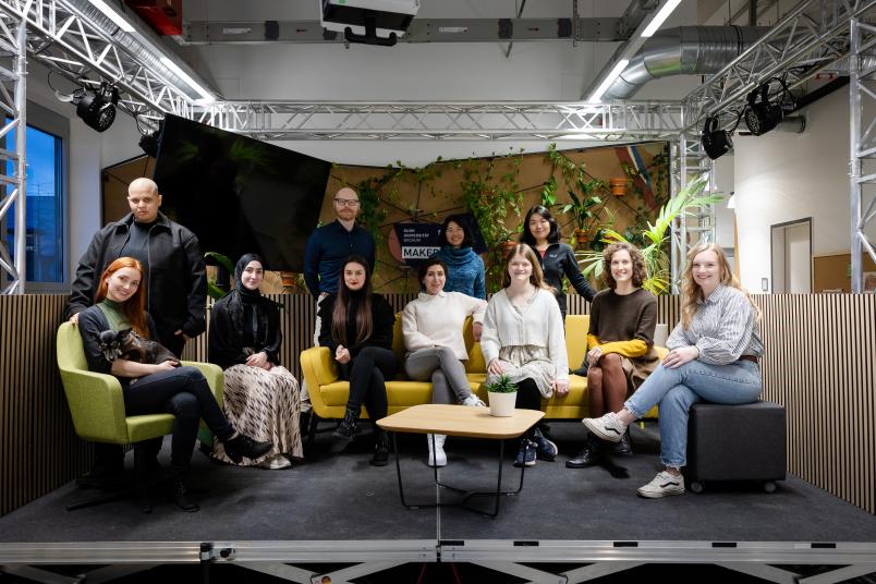 Das Gruppenfoto zeigt 11 junge Menschen. 7 junge Frauen sitzen auf einem gelben Sofa oder Stühlen im Vordergrund, zwei Männer und zwei Frauen stehen dahinter. Im Hintergrund sind Pflanzen und ein Monitor zu sehen.