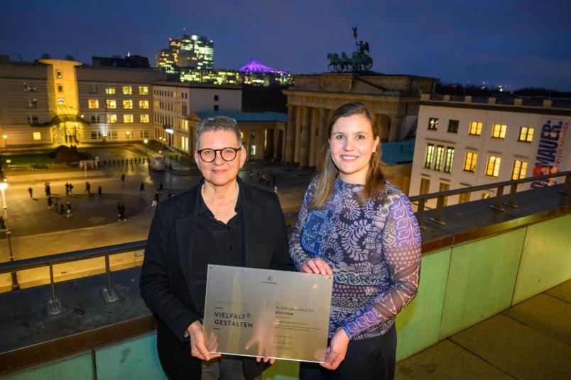 Das Foto zeigt zwei Frauen, die gemeinsam eine Urkunde festhalten. Die Frau links hat kurze Haare, trägt Brille und einen schwarzen Blazer; die Frau auf der rechten Seite hat mittellange blonde Haare und trägt ein blau-gemustertes Oberteil. Sie stehen auf einem Balkon draußen im Dunkeln. Im Hintergrund kann man das Brandenburger Tor sehen.