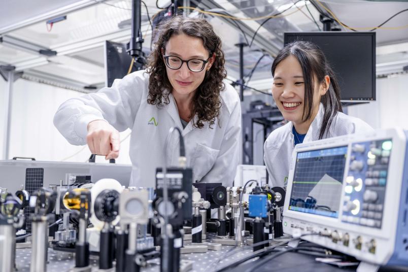 Das Bild zeigt zwei junge Forscherinnen in weißen Laborkitteln, die auf ein Experiment schauen und angeregt auf etwas zeigen.