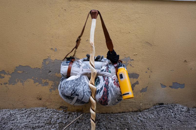 Wanderstab lehnt an einer Wand. Daran hängen ein Gepäckbeutel und eine Wasserflasche.