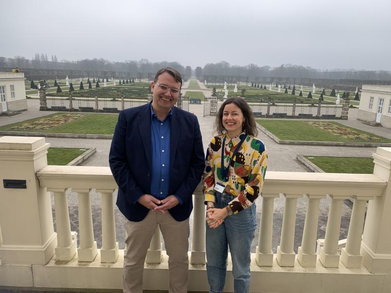 Ein Mann und eine Frau stehen nebeneinander; im Hintergrund sieht man einen Schlossgarten mit rechteckigen Rasenflächen, kleinen Tannen, Mauern und weißen Statuen. Der Mann trägt kurze schwarze Haare, ein blaues Hemd, ein dunkles Jackett und eine helle Hose. Die Frau steht rechts und trägt braune schulterlange Haare, eine bunt-getupfte Bluse und Jeans.