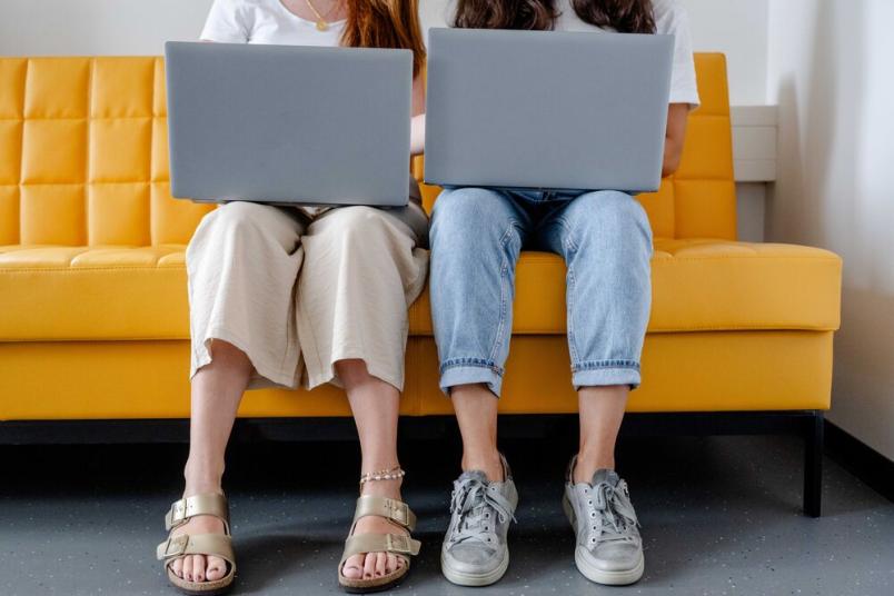 Zwei Menschen sitzen nebeneinander auf einer gelben Bank. Sie haben Laptops auf dem Schoß. 