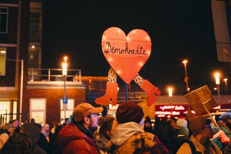 Ein Schild wird während einer Demo hochgehalten. Es ist Nacht. Das Pappschild ist in Herzform und trägt die Aufschrift Demokratie.
