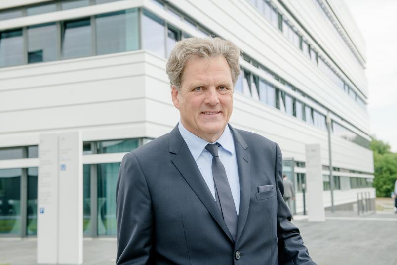 Porträt von Mann mit grauem Haar. Er trägt ein Jackett. Im Anzug sieht man ein Forschungsgebäude.