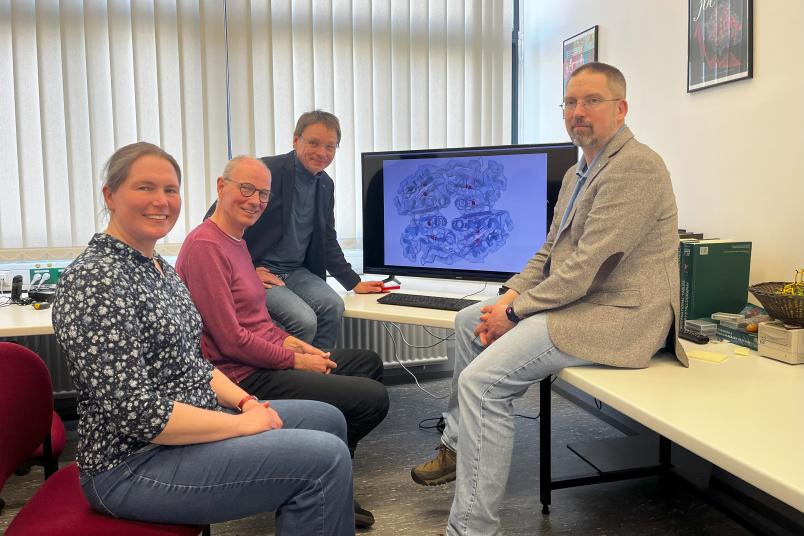 Vier Personen sitzen um einen großen Monitor herum, auf dem mehrere helixförmige Strukturen zu sehen sind.