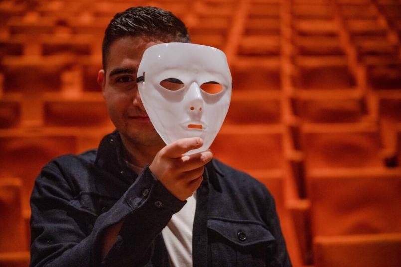 Eine Person hält eine weiße Maske vor das Gesicht, im Hintergrund rote Theatersitze.