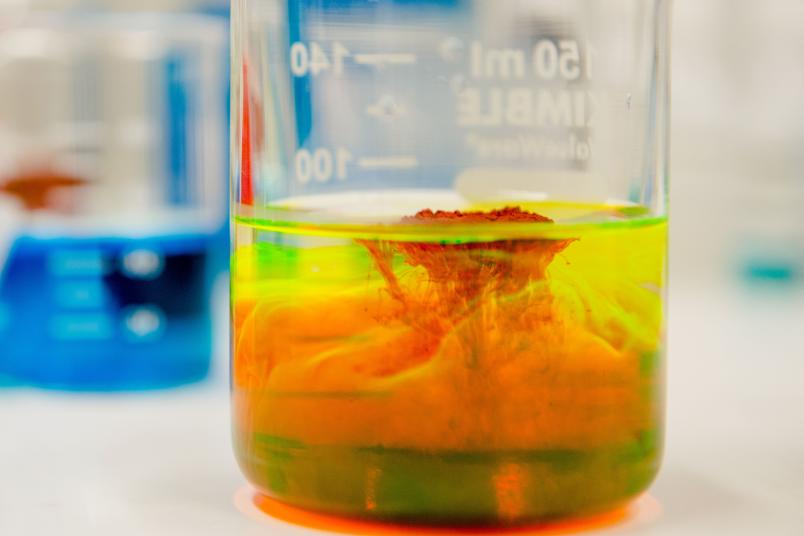 Eine rotes Pulver Flüssigkeit löst sich in einem Glasgefäß in einer gelben Flüssigkeit