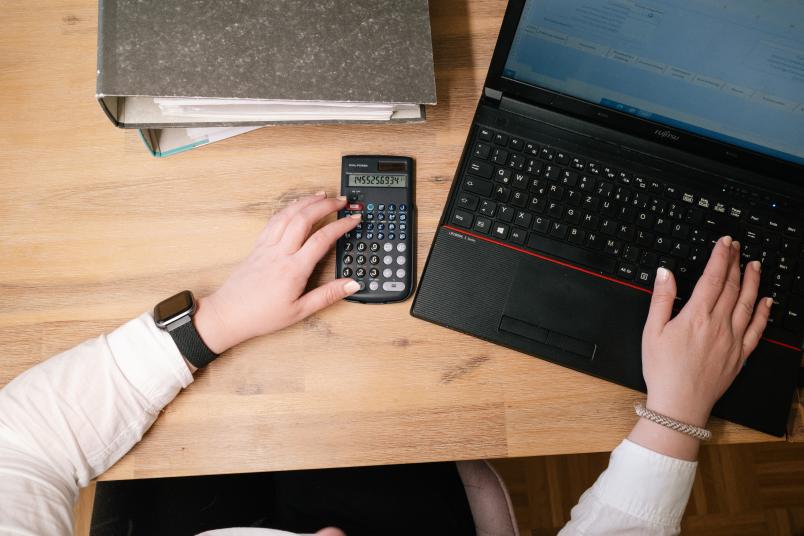 Draufsicht auf einen Schreibtisch, auf dem ein Laptop, ein Aktenordner und ein Taschenrechner zu sehen sind.