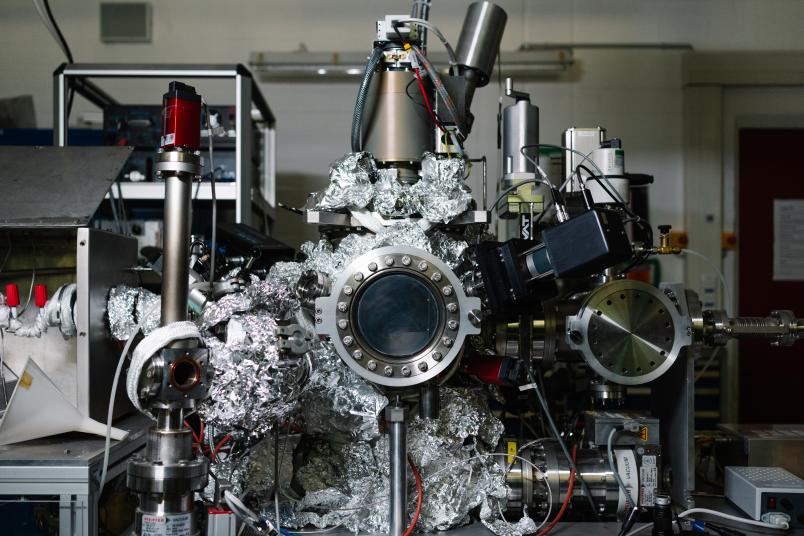 Ein metallischer Versuchsaufbau mit Bullauge, von dem einige Verbindungen mit Alufolie ummantelt sind