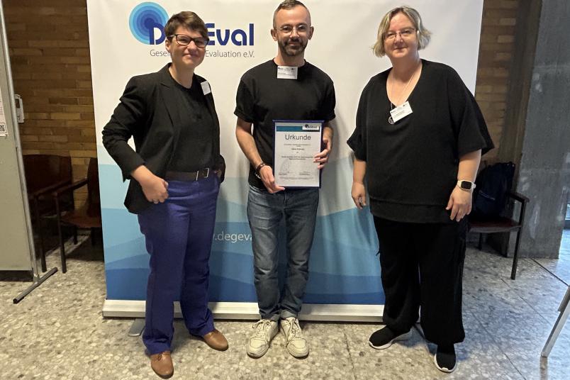 Gruppenbild, links und rechts jeweils eine Frau, in der Mitte ein Mann mit Urkunde