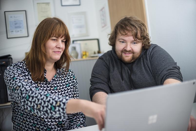 Eine langhaarige Frau und ein langhaariger Mann sprechen vor einem Laptop miteinander