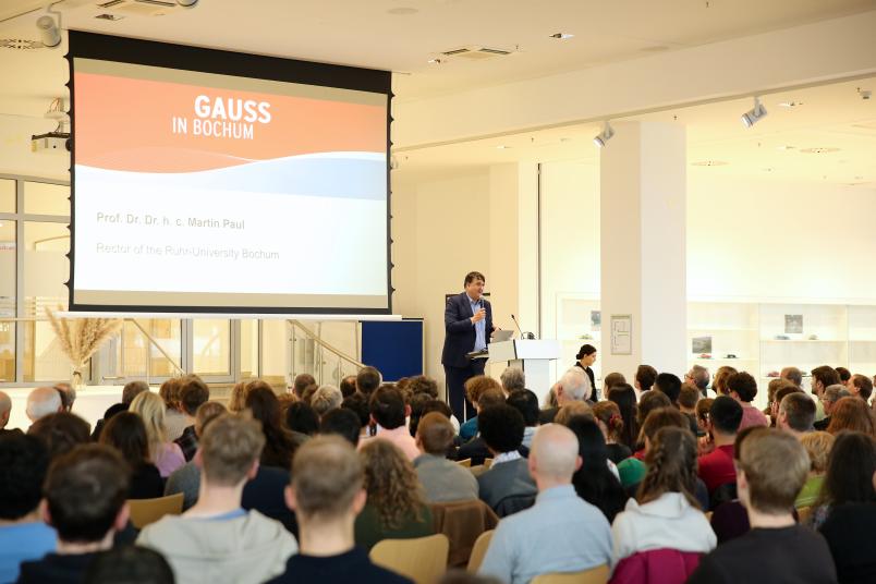 Rektor Martin Paul an einem Rednerpult in einem vollen Raum, dahinter eine Leinwand mit der Aufschrift "Gauss in Bochum"