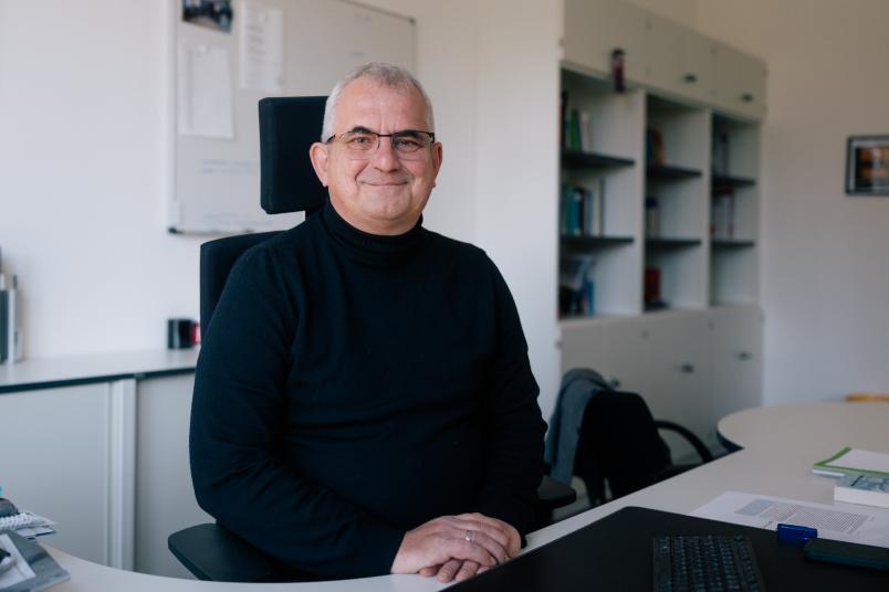 Mann mit schwarzem Rolli und grauen Haaren sitzt an einem Schreibtisch und lächelt in die Kamera.