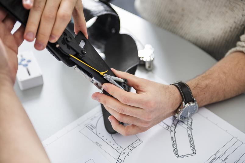 Zwei Hände halten einen Orthesen-Prototypen, darunter liegt eine Bauskizze auf einem Tisch.