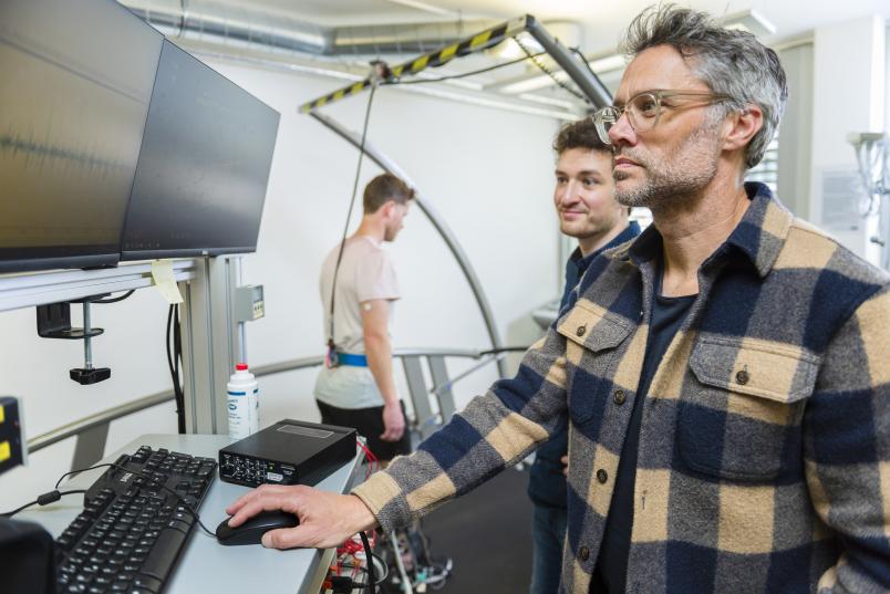Ein Mann geht im Hintergrund auf einem Laufband. Im Vordergrund sind zwei Männer zu sehen, die auf einen Monitor schauen. 