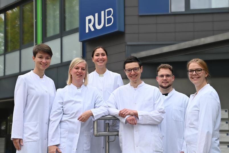 Ein Gruppenfoto von sechs Personen in weißen Kitteln vor dem RUB-Würfel