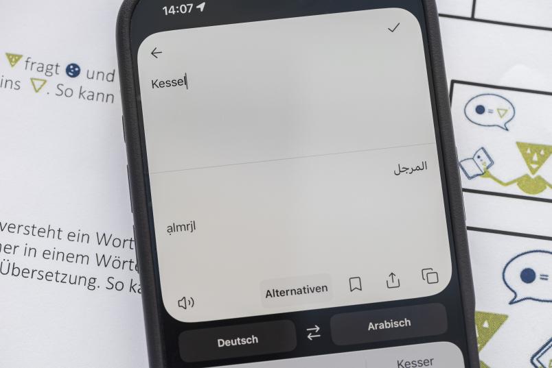 Eine Übersetzungsapp auf einem Handy zeigt das Wort "Kessel" in arabischer Schrift.