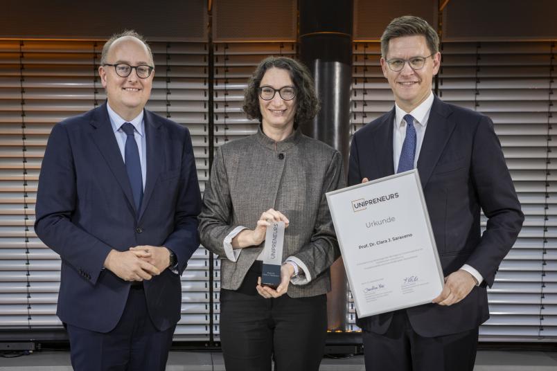 Preisträgerin Clara Saraceno mit den Staatssekretären Matthias Hauer (BMFTR, links) und Johannes Steiniger (BMWE).