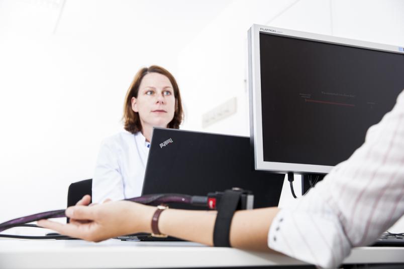 Eine Frau sitzt an einem Monitor, ihr gegenüber eine Versuchsperson mit einer Thermode am Arm