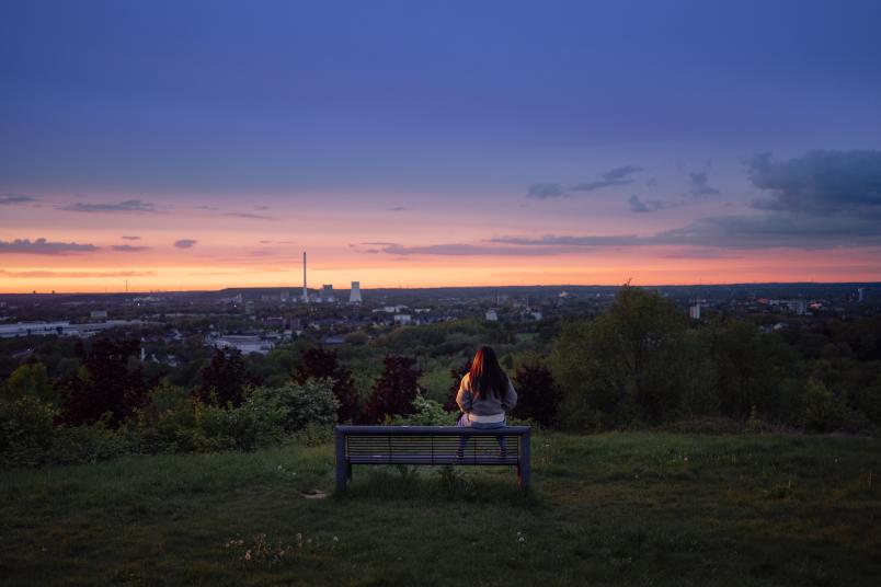 Eine einzelne Person sitzt auf einer Anhöhe auf einer Bank und schaut über das Ruhrgebiet.