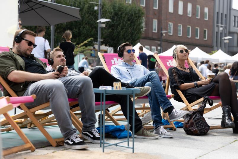 Junge Leute sitzen in pinken Liegestühlen auf dem Rathausvorplatz in Bochum und haben Kopfhörer auf.