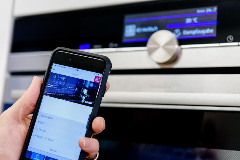 Eine Hand hält ein Smartphone vor einen Ofen. Auf dem Display sind diverse Einstellmöglichkeiten für das Gerät zu sehen.