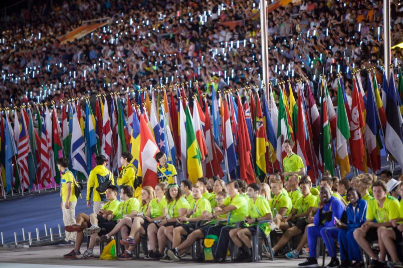 Viele Athletinnen und Athleten sitzen vor einem Flaggenmeer in einem Stadion