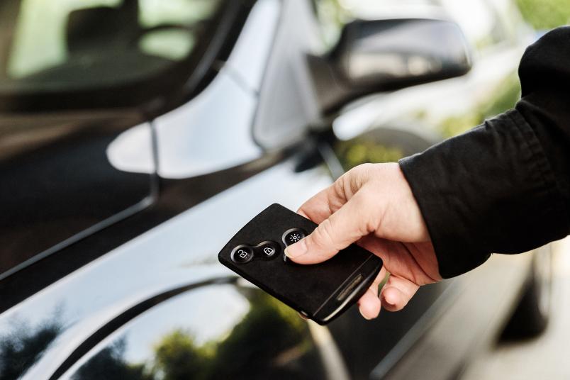 Eine Hand betätigt einen elektronischen Autoschlüssel. Im Hintergrund ist ein Auto zu sehen.