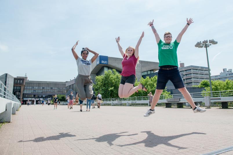 Drei Personen springen mit ausgestreckten Armen, fröhlich auf dem Campus in die Luft.