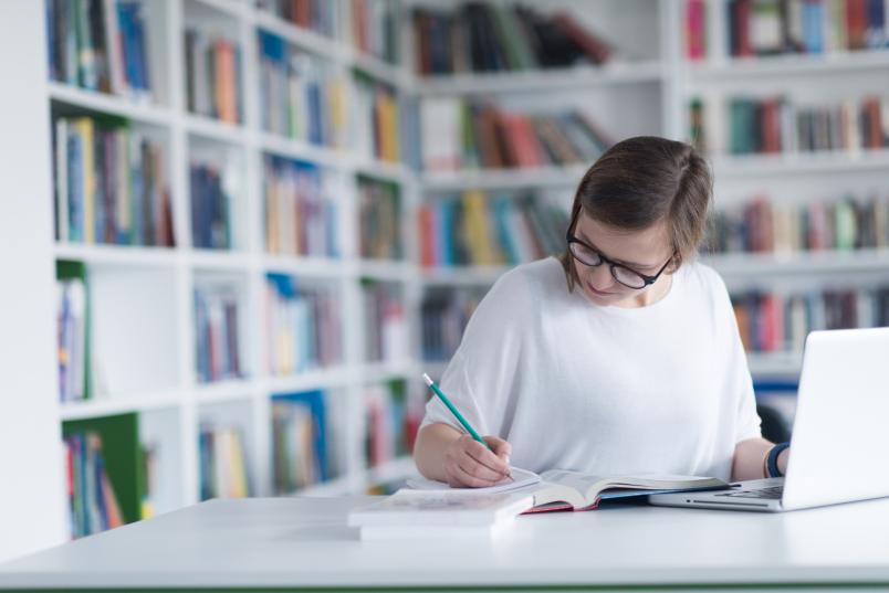 Frau mit Laptop und Büchern macht Notizen in einer Bibliothek