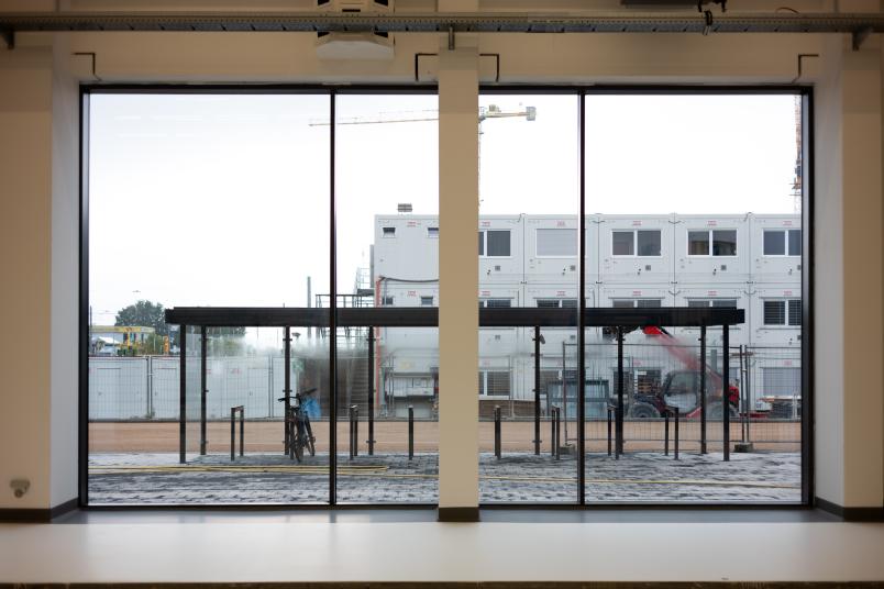 Blick durch große Fensterfronten auf Fahrradständer, die überdacht sind. Baucontainer im Hintergrund.