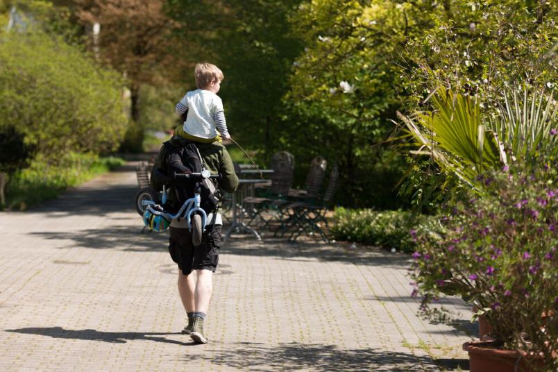 Person mit einem Kind auf den Schultern und einem Kinderfahrrad in der Hand spaziert durch den botanischen Garten der RUB.