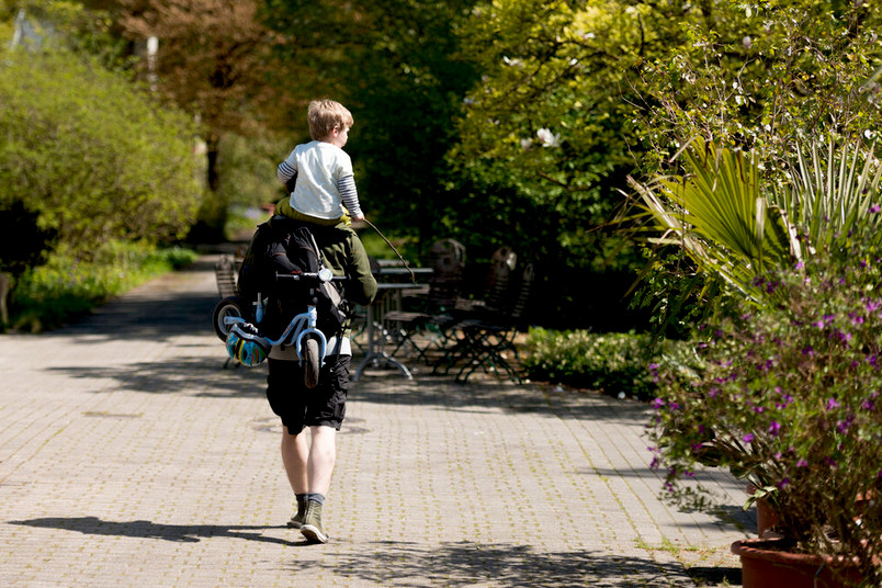 Person mit einem Kind auf den Schultern und einem Kinderfahrrad in der Hand spaziert durch den botanischen Garten der RUB.