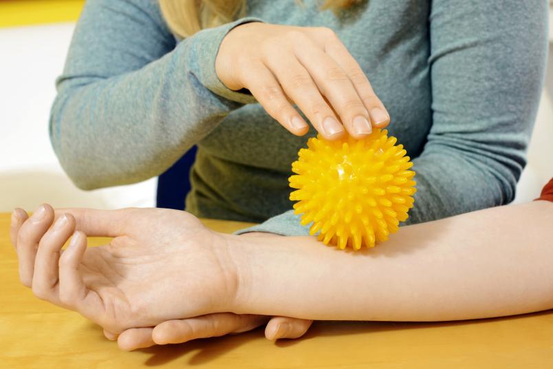 Eine Ergotherapeutin fährt mit einem Igelball über den Arm eines Patienten.