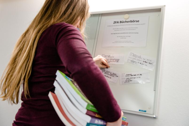 Eine Frau mit Büchern unter dem Arm hängt einen Zettel in eine Vitrine.