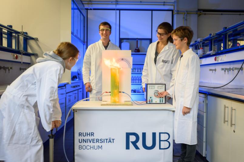 Zwei Kinder und zwei Forscher beobachten, wie eine Flamme aus einem Plexiglaszylinder schießt.