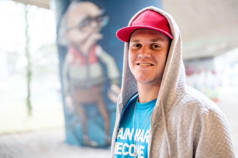 Ein junger Mann mit Kapuze über der Cappie steht vor einem Graffiti