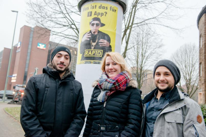 Dozentin Katharina Friedrichs mit zwei Studenten vor dem Plakat an einer Litfaßsäule