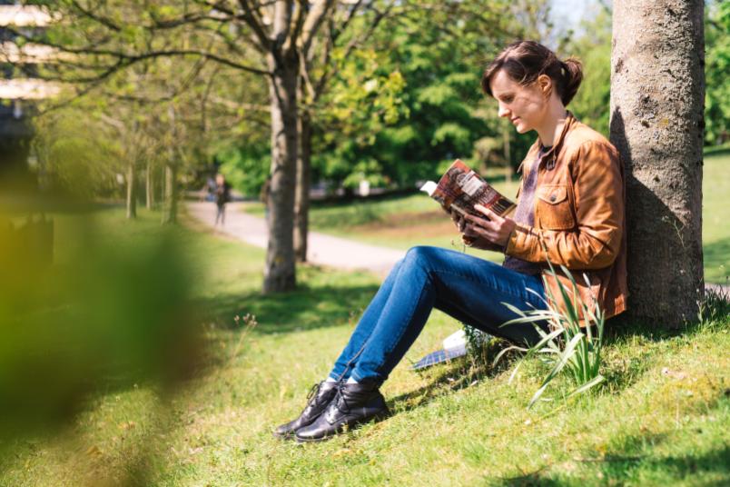 Porträt einer etwa 30-jährigen Frau, die auf der Wiese sitzt und ein Buch liest