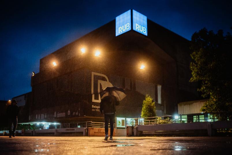 Person bei Nacht mit aufgespanntem Regenschirm, im Regen vor einem RUB Gebäude.