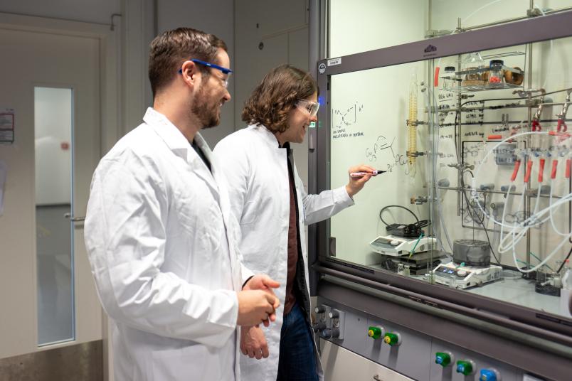 Zwei Forscher in Kitteln schreiben chemische Formeln an eine Glasscheibe.
