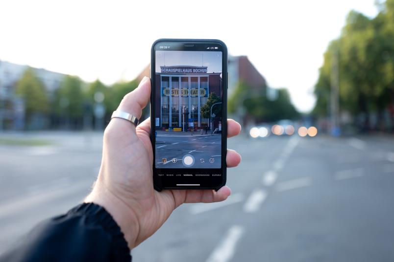 Eine Hand hält ein Smartphone, das ein Theatergebäude zeigt.