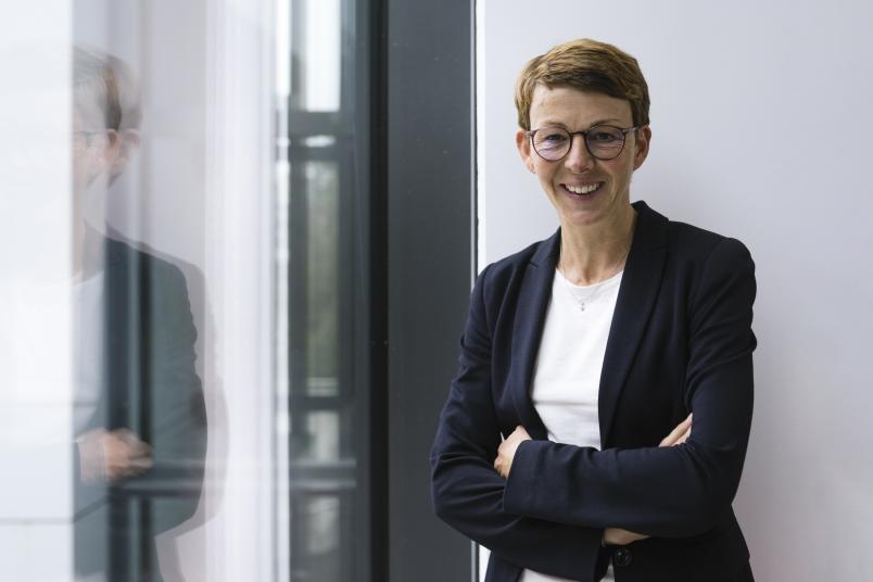 Eine Frau in schwarzem Blazer steht lachend an einem Fenster