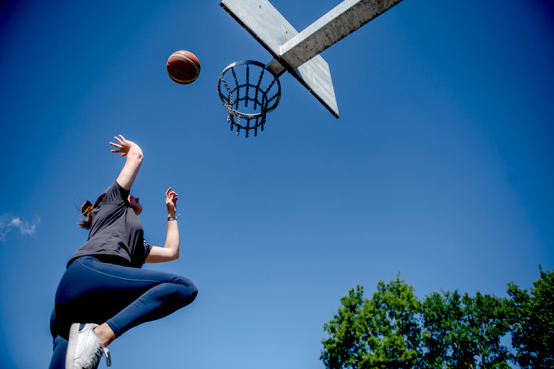 Eine Frau spingt zu einem Basketballkorb hoch und wirft einen Basketball. Von unten fotografiert vor blauem Himmel.