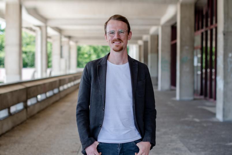 Portrait von Jonas Leschke. Er steht auf dem Campus und blickt lächelnd in die Kamera.